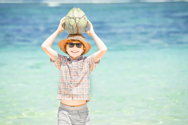 Счастливый мальчик, живущий на прекрасном пляже океана в шляпе и солнцезащитных очках с кокосом в руках — стоковое фото