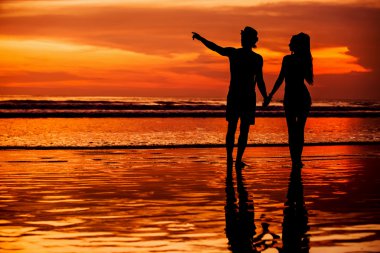 Silhouettes of young couple in love staing on the beach with beautiful red sunset as background clipart