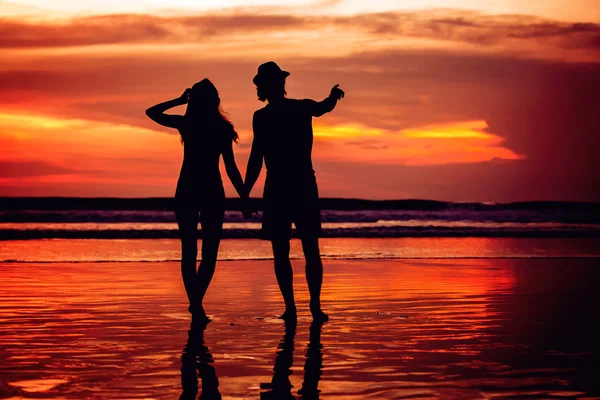 Silhouettes of young couple in love staing on the beach with beautiful red sunset as background — Stock Photo, Image