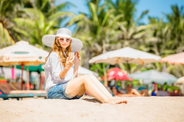 Trzymać młoda kobieta siedzi na plaży w słoneczny dzień z telefonem w dłoni — Zdjęcie stockowe