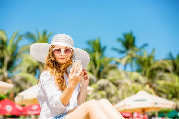 Trzymać młoda kobieta siedzi na plaży w słoneczny dzień z telefonem w dłoni — Zdjęcie stockowe