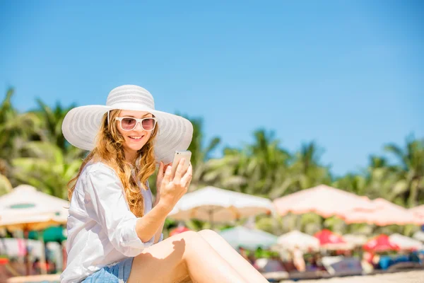 Beautiful young woman sitting on the beach at sunny day with phone in her hand — стоковое фото