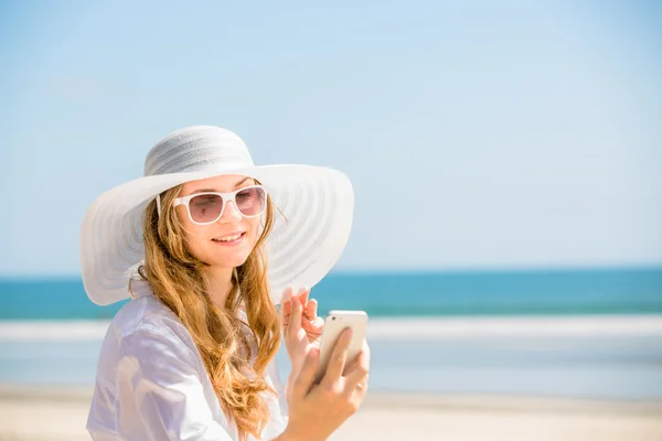 Beautifil young woman sitting on the beach at sunny day with phone in her hand — Stock Photo, Image
