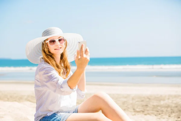 Beautifil joven sentada en la playa en un día soleado con teléfono en la mano —  Fotos de Stock