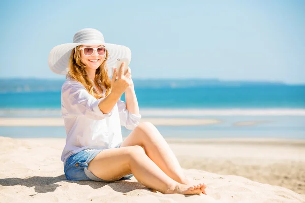 Beautiful young woman sitting on the beach at sunny day with phone in her hand — стоковое фото