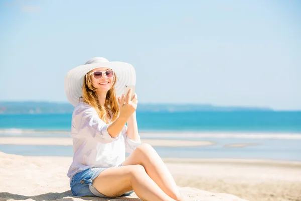 Trzymać młoda kobieta siedzi na plaży w słoneczny dzień z telefonem w dłoni — Zdjęcie stockowe