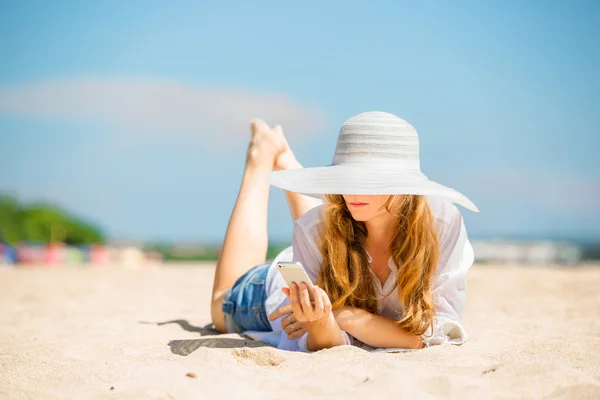 Trzymać młoda kobieta, leżąc na plaży w słoneczny dzień z telefonem w dłoni — Zdjęcie stockowe