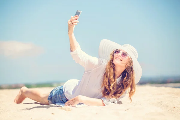 Beautifil young woman lying on the beach at sunny day and taking selfie with phone — Stock Photo, Image