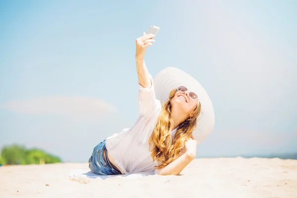 Beautifil young woman lying on the beach at sunny day and taking selfie with phone — Stock Photo, Image