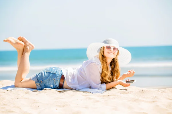 Beautifil joven mujer acostada en la playa en un día soleado con el teléfono en la mano —  Fotos de Stock