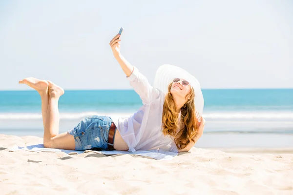 Beautiful young woman lying on the beach at sunny day and taking selfie with phone — стоковое фото