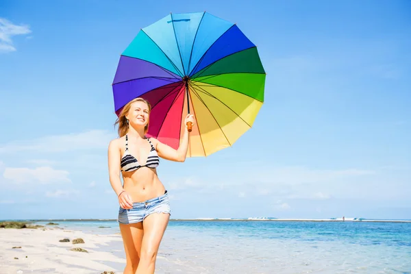 Young woman in blue shorts with colourful rainbow umbrella walking on beach — Stock Photo, Image