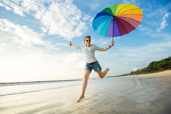 Jovem alegre com guarda-chuva arco-íris se divertindo na praia antes do pôr do sol. Viagens, feriados, férias, conceito de estilo de vida saudável — Fotografia de Stock