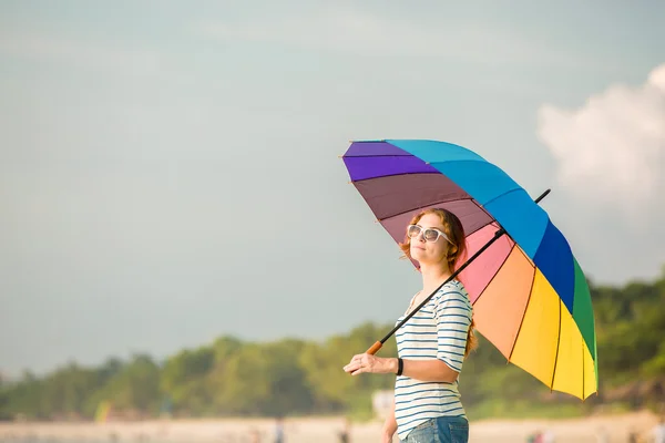 Young caucasian woman wearing sunglasses with colourful rainbow umbrella looking at ocean on the beach before sunset. Travel, holidays, vacation, healthy lifestyle concept — Stock Photo, Image