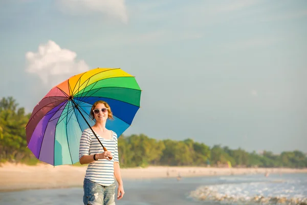Καυκάσιος κοπέλα φορώντας γυαλιά ηλίου με πολύχρωμο ουράνιο τόξο ομπρέλα κοιτάζοντας ωκεανό στην παραλία πριν την δύση του ηλίου. ταξίδια, διακοπές, διακοπές, έννοια του υγιεινού τρόπου ζωής — Φωτογραφία Αρχείου