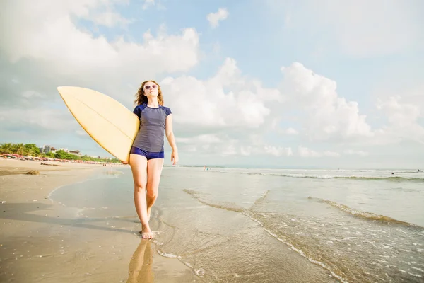 Beautiful young caucasian girl in rushwest and sunglasses with yellow surfboard at legian beach, Bali. Lyfestyle, leisure, sport, vacation, happines concept — Stock Photo, Image