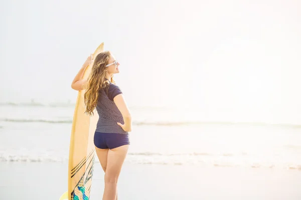 Beautiful young surfer girl stays on the beach at sunset with yellow surfboard. Healthy lifestyle, leisure, travel, holiday and sport concept, copyspace — Stock Photo, Image