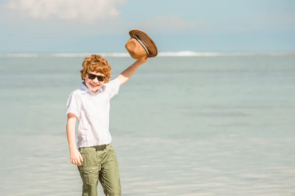 Gelukkig red haired kind jongen dragen van een hipster zonnebril zwaaien hoed genieten van prachtige zonnige zomerdag blijft op het strand in ondiep water — Stockfoto