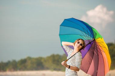 Attractive caucasian woman wearing sunglasses holding colourful rainbow umbrella looking at ocean on the beach before sunset. Travel, holidays, vacation, healthy lifestyle concept clipart