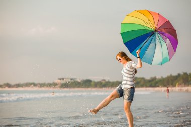 Cheerful young girl with rainbow umbrella having fun on the beach before sunset. Travel, holidays, vacation, healthy lifestyle concept clipart