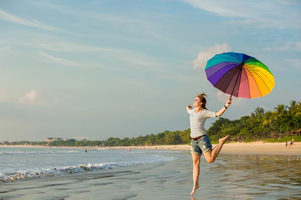 Cheerful young girl with rainbow umbrella having fun on the beach before sunset. Travel, holidays, vacation, healthy lifestyle concept — Stock Photo, Image