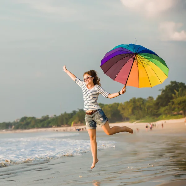 Cheerful young girl with rainbow umbrella having fun on the beach before sunset. Travel, holidays, vacation, healthy lifestyle concept — Stock Photo, Image