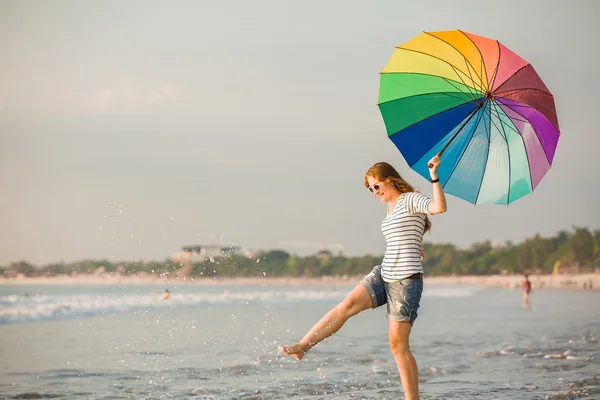 Εύθυμη νεαρή κοπέλα με ομπρέλα ουράνιο τόξο τη διασκέδαση στην παραλία πριν την δύση του ηλίου. ταξίδια, διακοπές, διακοπές, έννοια του υγιεινού τρόπου ζωής — Φωτογραφία Αρχείου