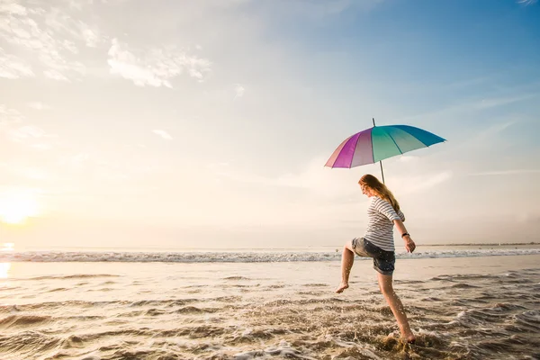 Vidám fiatal lány szivárvány esernyő szórakozás a strandon naplemente előtt. utazás, üdülés, nyaralás, egészséges életmód fogalma — Stock Fotó