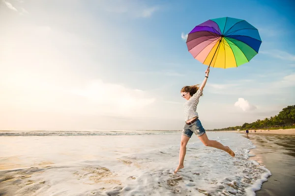 Glad ung flicka med rainbow paraply att ha kul på stranden före solnedgången. Res, semester, semester, hälsosam livsstilskoncept — Stockfoto