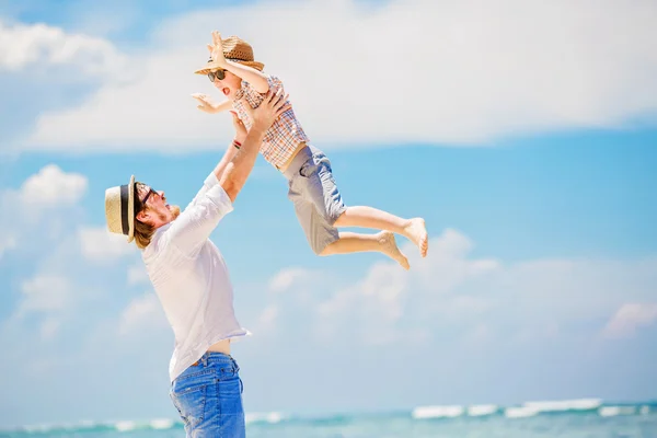 Young happy father holding little son in his arms putting him up standing barefoot at the beach with ocean and beautiful clouds on background — Stock Photo, Image