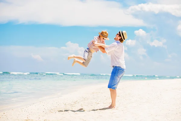 Young happy father holding little son in his arms putting him up standing barefoot at the beach with ocean and beautiful clouds on background — 스톡 사진