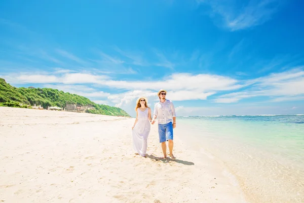 Jovem casal feliz em chapéus e óculos de sol caminhar na praia tropical com areia branca contra bela vista sobre o fundo — Fotografia de Stock