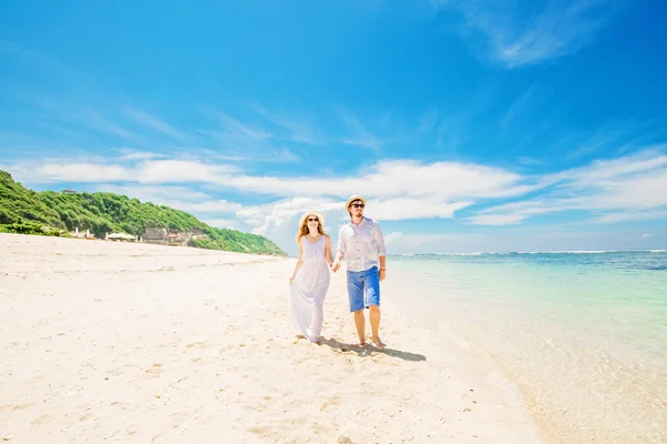 Happy young couple in hats and sunglasses walk at tropical beach with white sand against beautiful view on background — Zdjęcie stockowe