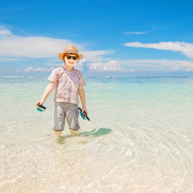 Happy kid boy wearing summer hat and hipster sunglasses walks in shallow water enjoying sunny day