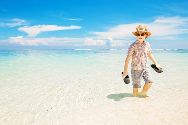 Happy kid boy wearing summer hat and hipster sunglasses walks in shallow water enjoying sunny day — 스톡 사진