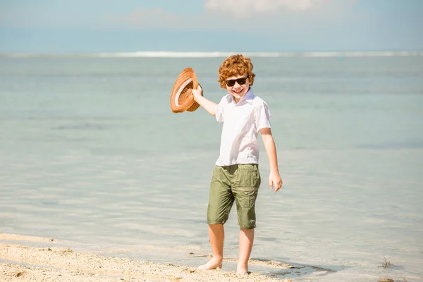 Gelukkig red haired kind jongen dragen van een hipster zonnebril zwaaien hoed genieten van prachtige zonnige zomerdag blijft op het strand in ondiep water — Stockfoto