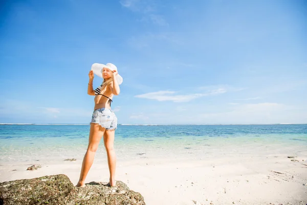 Young skinny caucasian girl with tan stays on the rock facing ocean, turning to camera against blue sky and pure water. Travel, vacation, paradise concept, copyspace — Stock Photo, Image