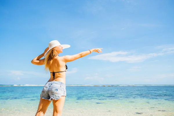 Skinny caucasian tanned woman stays on the rock against blue sky and pure ocean water, pointing with her hand. Travel, vacation, paradise concept, copyspace — Stockfoto