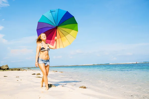 Mujer joven en pantalones cortos azules con colorido paraguas de arco iris caminando en la playa — Foto de Stock