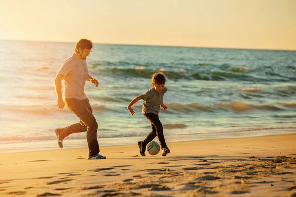 Happy father and son play soccer or football on the beach in sunset light — Zdjęcie stockowe