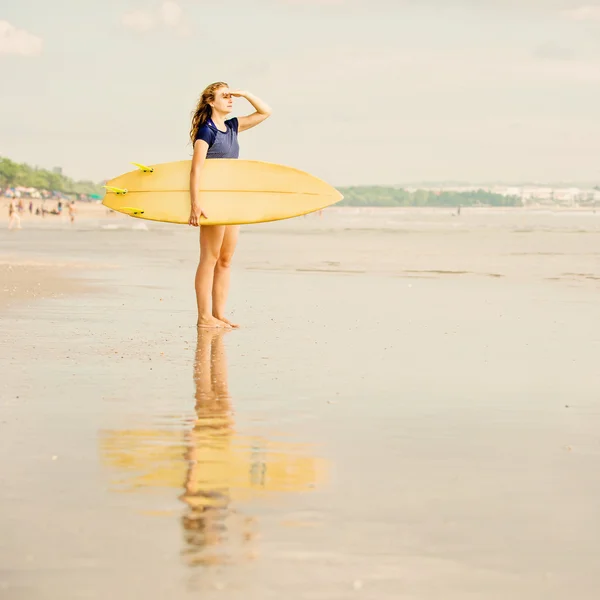 Beautiful sexy surfer girl on the beach at sunset looking for waves from shore with surfboard in her hands — Stock Photo, Image