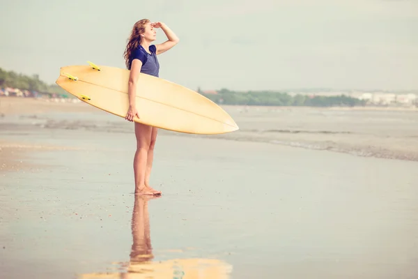 Beautiful sexy surfer girl on the beach at sunset looking for waves from shore with surfboard in her hands — Stock Photo, Image