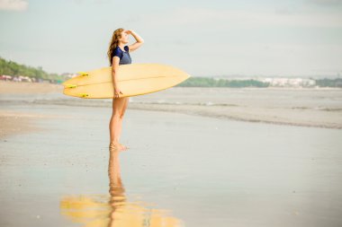 Beautiful sexy surfer girl on the beach at sunset looking for waves from shore with surfboard in her hands