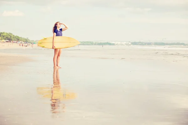 Beautiful sexy surfer girl on the beach at sunset looking for waves from shore with surfboard in her hands — Zdjęcie stockowe