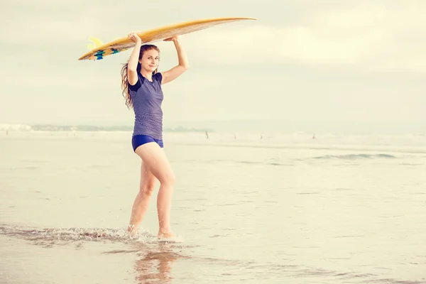 Hermosa chica surfista sexy en la playa al atardecer camina a lo largo de la orilla del océano, tabla de surf amarillo en sus manos. Vida sana, concepto deportivo con copyspace —  Fotos de Stock