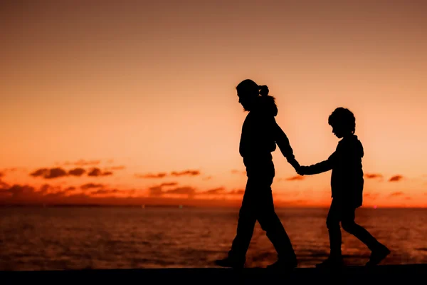 Silhouette einer jungen Mutter und ihres Sohnes, die an einem Sommertag an einem Steg vor einem Sonnenuntergang am Himmel spazieren gehen — Stockfoto