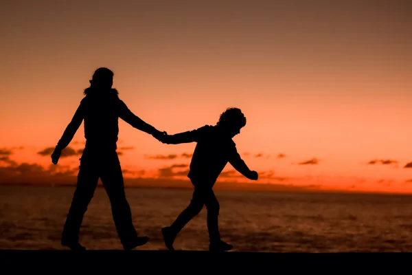 Silueta de una joven madre y su hijo caminando por un muelle frente a una puesta de sol en el cielo en un día de verano — Foto de Stock