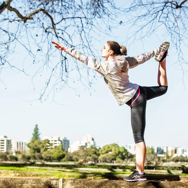 Giovane donna che si estende sulla panchina prima di fare jogging nella natura autunnale con la città sullo sfondo — Foto Stock