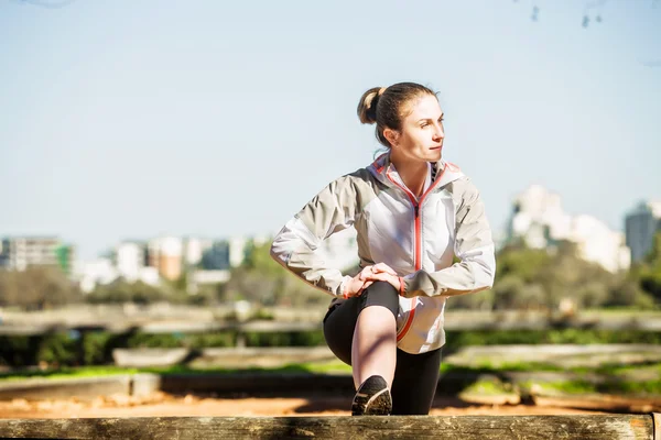Young fit girl stretching out on bench before exercising in autumn pak with city at background — Stock Photo, Image