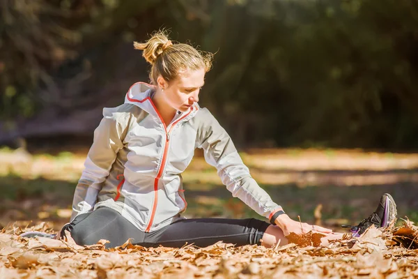 Joven mujer deportiva en forma estirándose antes de trotar sentado en hojas amarillas de otoño — Foto de Stock